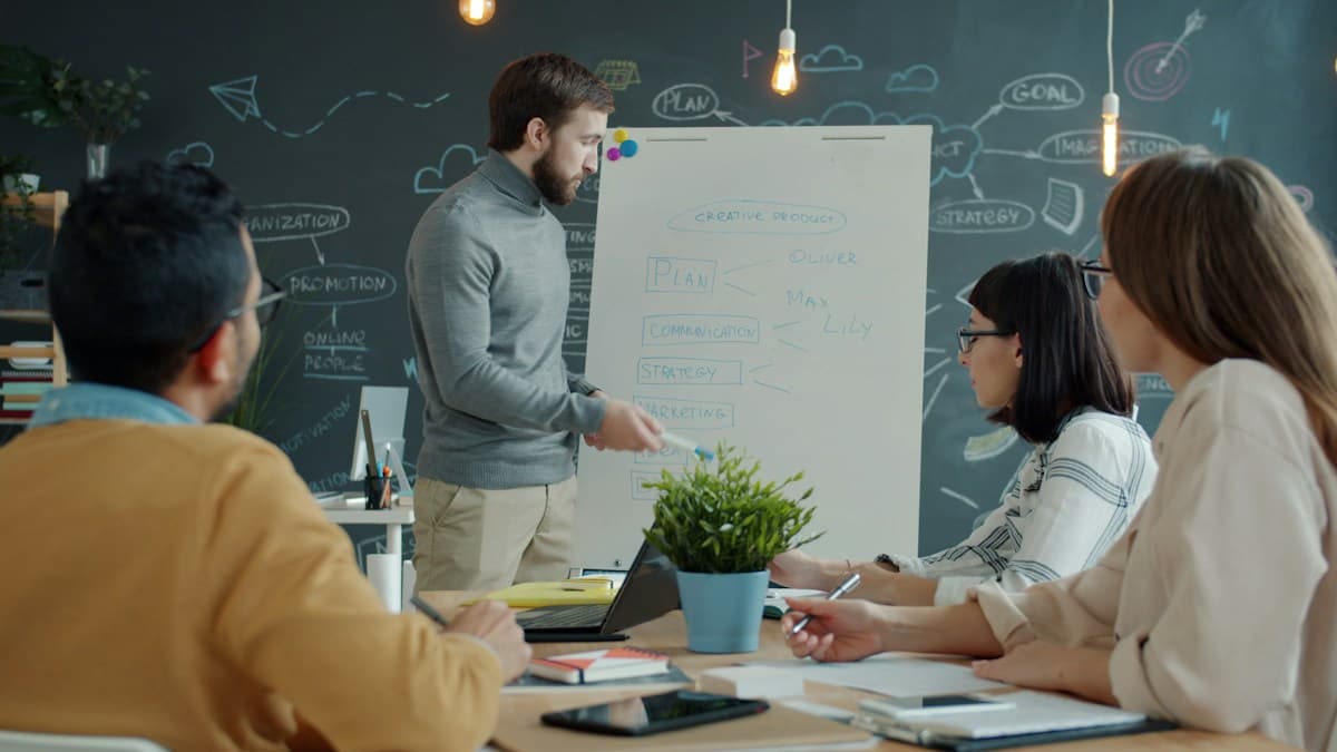 Un formador explica junto a una pizarra a un grupo sentado a la mesa, como en una sesión de formación ERPNext en sala