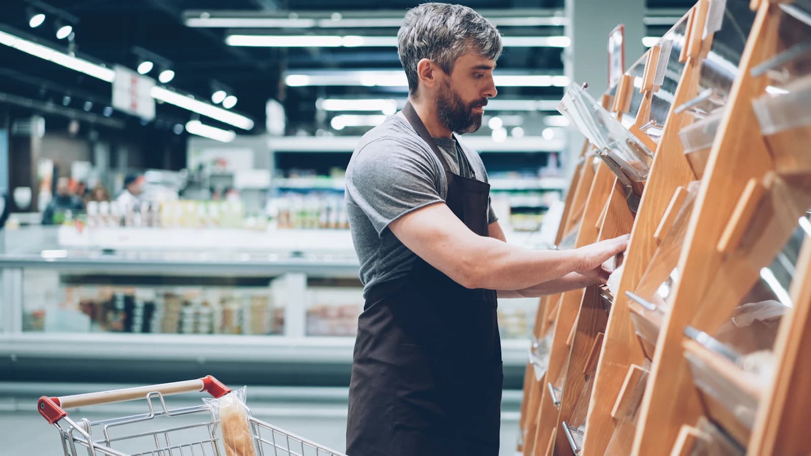 Dependiente con delantal reponiendo un expositor de madera en el lineal de una tienda de alimentación, el stock que mueve un comercio